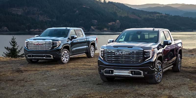 Two GMC Sierra pickup trucks parked beside a lakeside overlook with mountains in the background