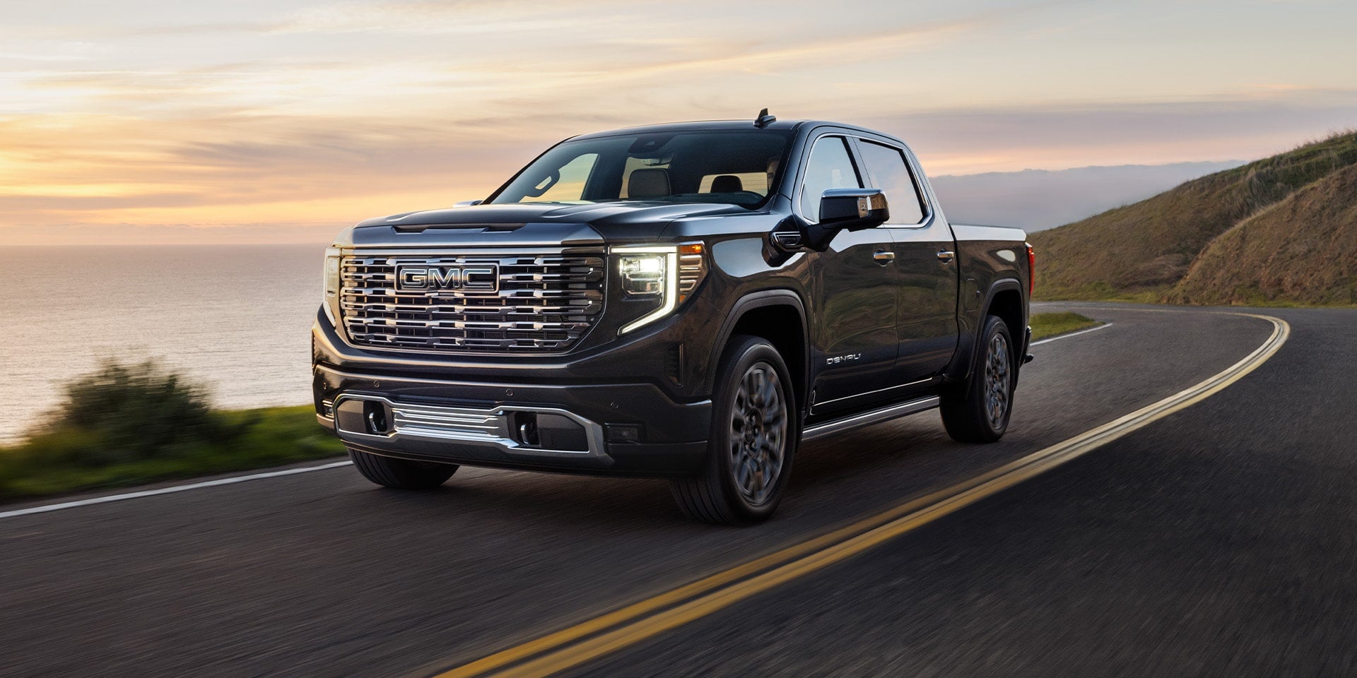 Black GMC pickup truck driving along a coastal highway at sunset, shown from a front three-quarter angle with ocean views and cliffs in the background