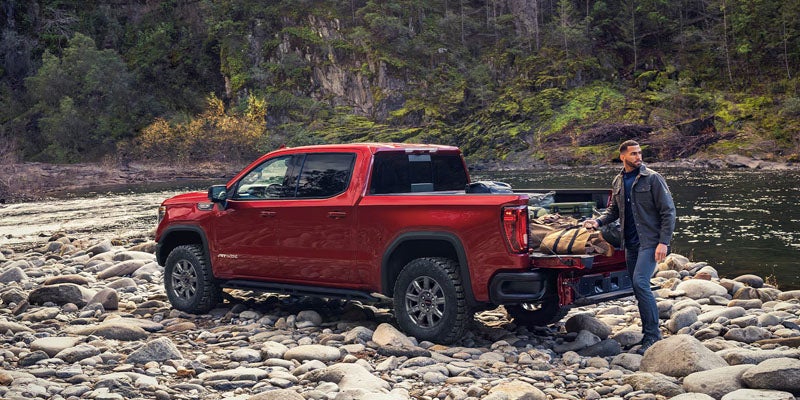 Red GMC Sierra pickup truck parked on a rocky riverbank with outdoor gear in the truck bed