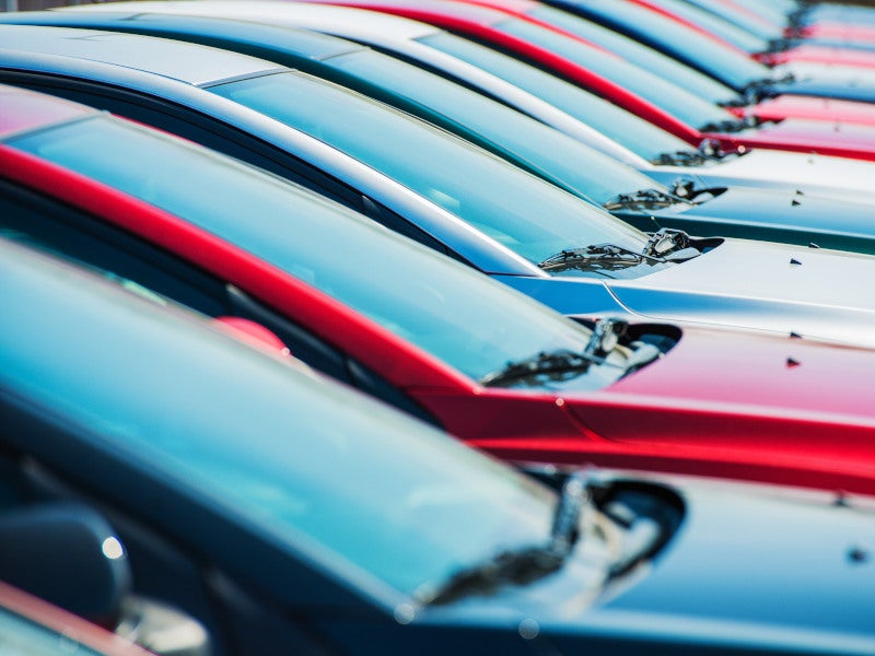 Diagonal view of a row of parked cars with red, blue, and silver roofs, showcasing windshields and sleek body lines in a dealership lot