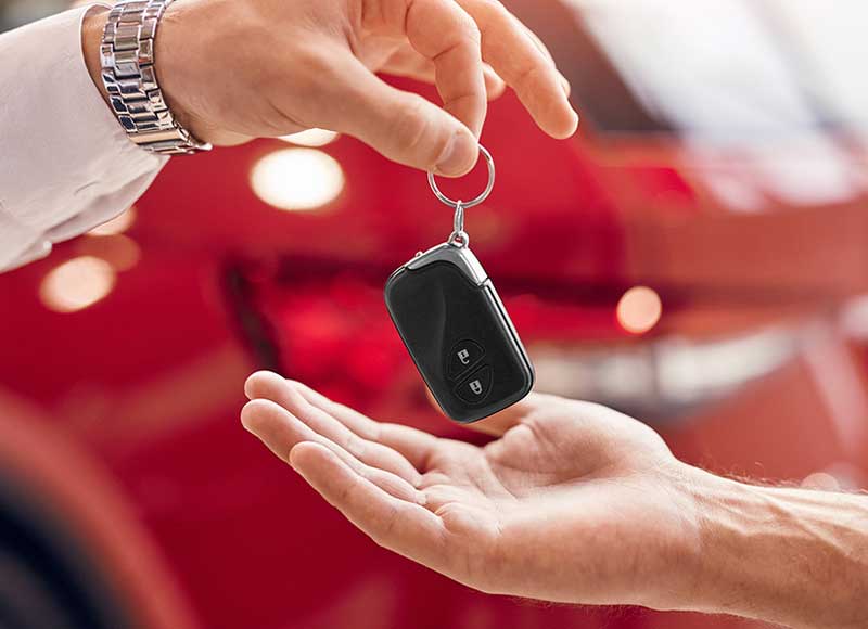 Close-up of a car key fob being handed from one person to another, with a red vehicle blurred in the background